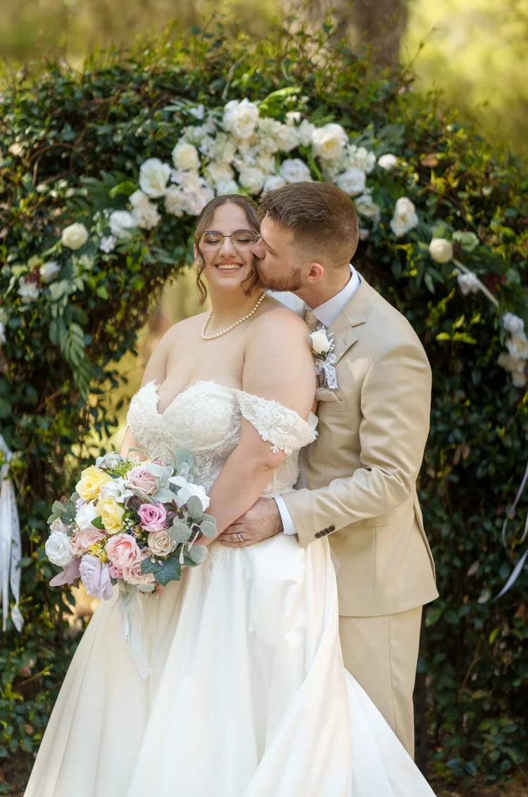 A groom kisses his bride's temple beneath a full floral and greenery arch, her pastel bouquet held at her side.
