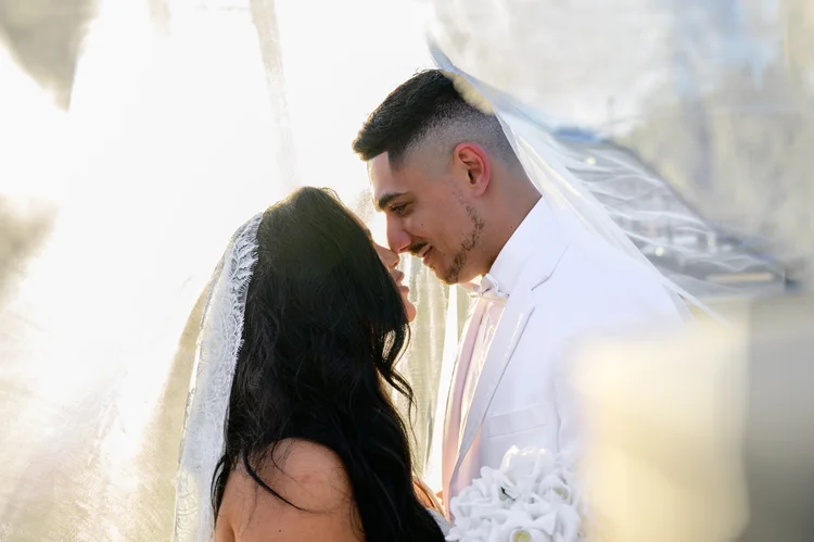 A bride and groom rest their foreheads together beneath her veil, backlit by warm afternoon sun.