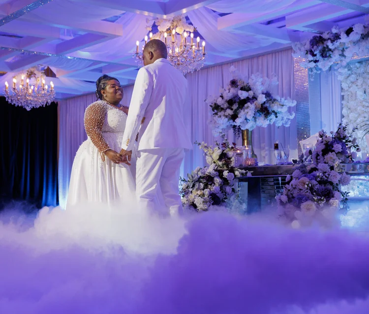 A couple share a first dance surrounded by low-lying fog on the dance floor, under violet lighting and crystal chandeliers.