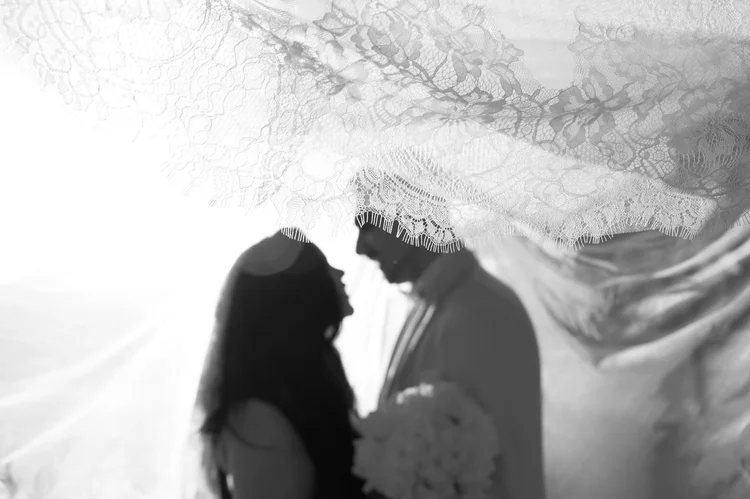 A black and white photograph of a bride and groom facing each other, silhouetted beneath the lace edge of her veil.