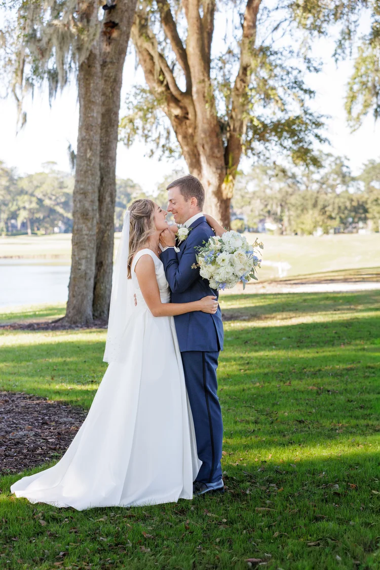 A bride and groom are standing outdoors near a large tree, smiling and gazing into each other's eyes during their wedding. The bride is wearing a white wedding dress with a veil, and the groom is dressed in a navy suit holding a bouquet of white and blue flowers.