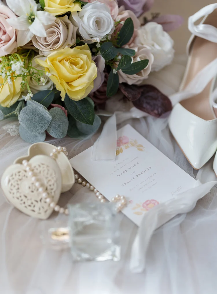 A flat-lay of wedding details: a bouquet of yellow, white and blush roses with eucalyptus, an invitation suite, the bride's shoes, and her jewellery arranged on soft tulle.
