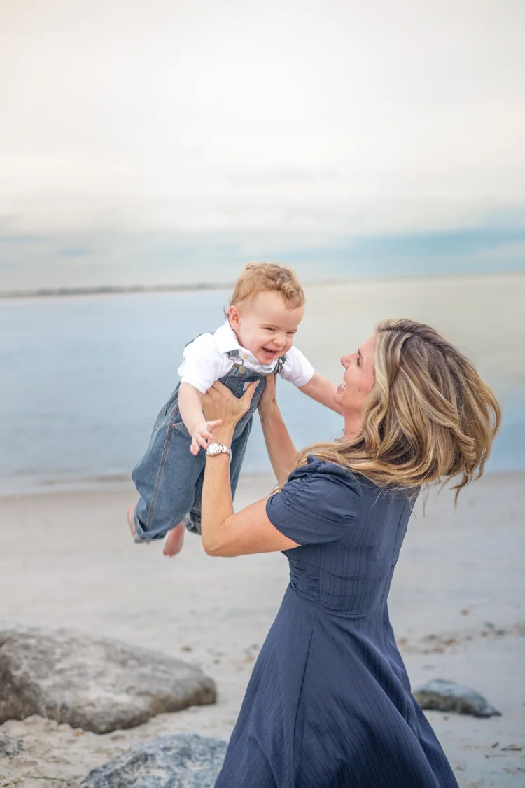 A mother in a navy dress lifts her laughing toddler into the air on the beach at sunset.