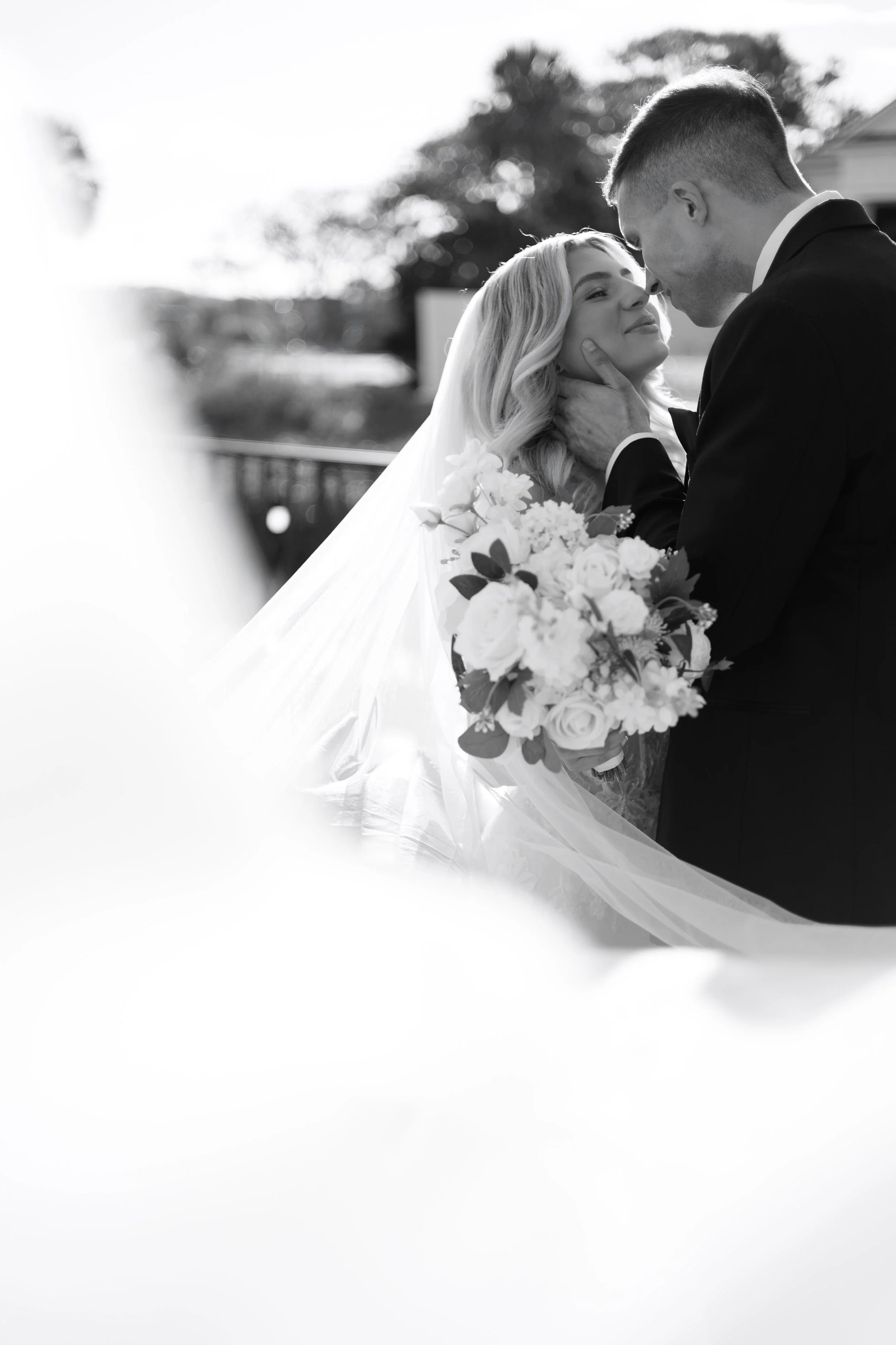 Black and white photo of a newlywed couple sharing an intimate moment outdoors; the bride is holding a bouquet of flowers, and they are gazing affectionate at each other.
