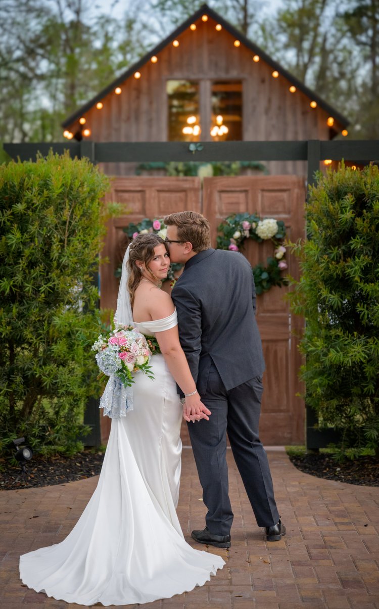 A bride and groom stand together in front of the lit doorway of a wooden barn, framed by greenery and a floral garland.