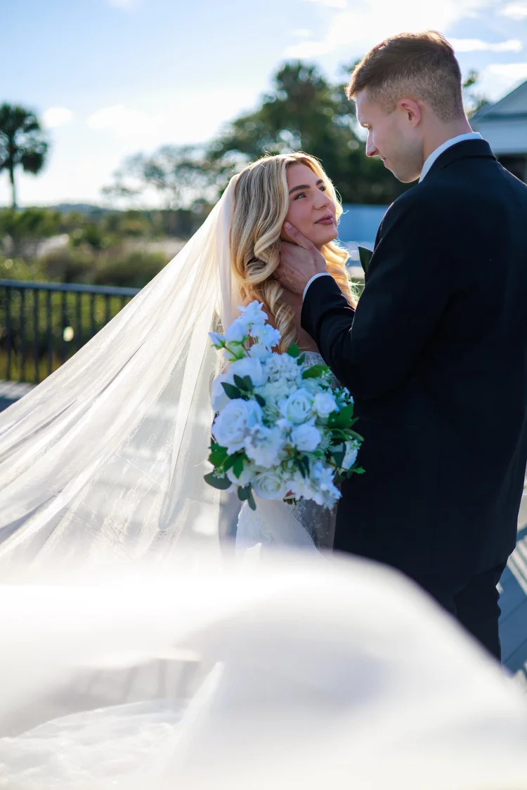 A bride's long veil catches the light as she and her groom stand close together on an outdoor terrace, her bouquet of white and pale blue flowers between them.