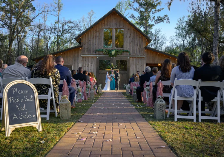 Guests seated on white chairs along a brick aisle facing a rustic wooden barn set for a ceremony, with a hand-lettered sign at the aisle entrance.