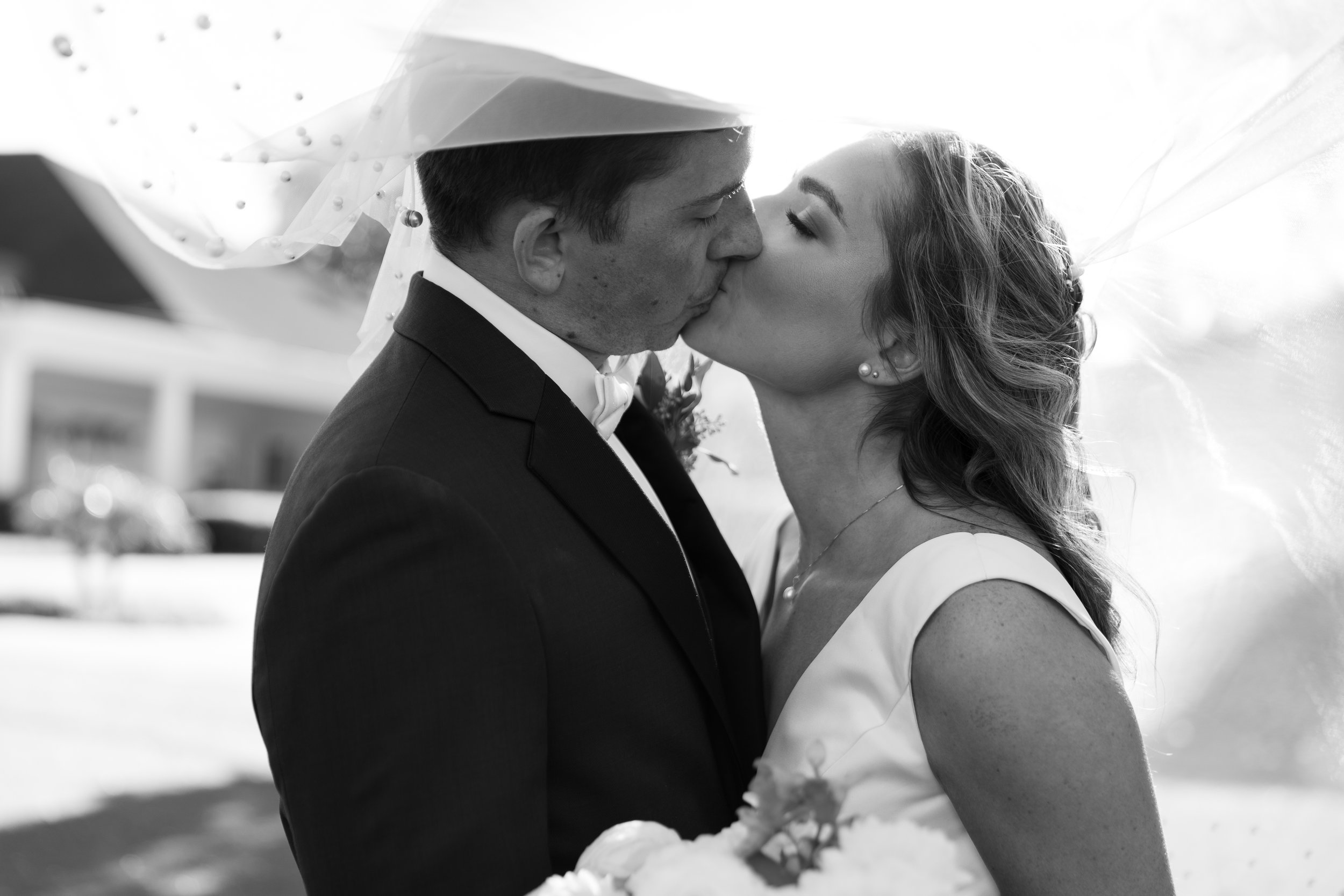 A black and white photo of a bride and groom sharing a kiss outdoors, with the bride holding a bouquet.