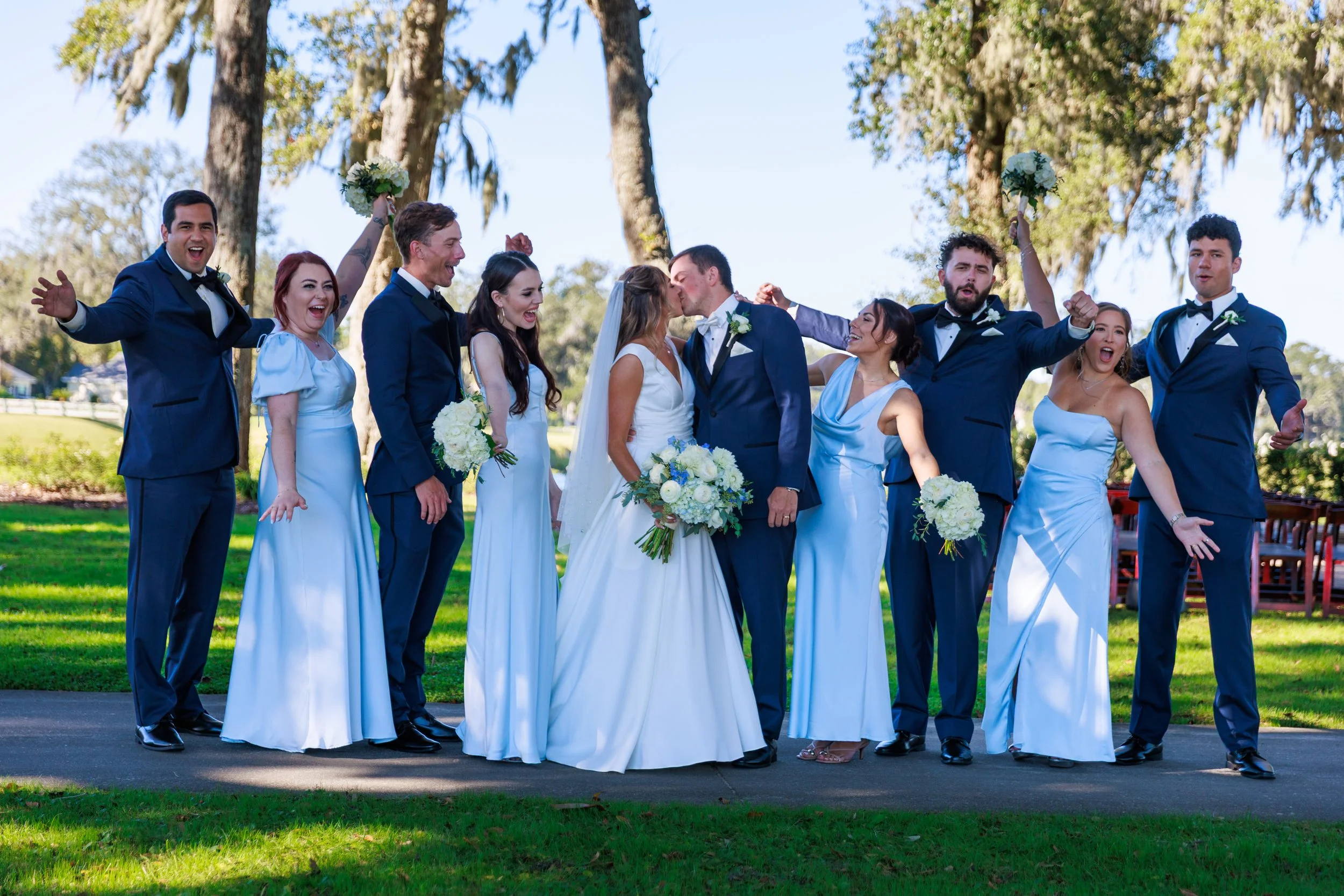 A wedding party outside in a park, with the bride and groom kissing surrounded by bridesmaids and groomsmen smiling and celebrating.