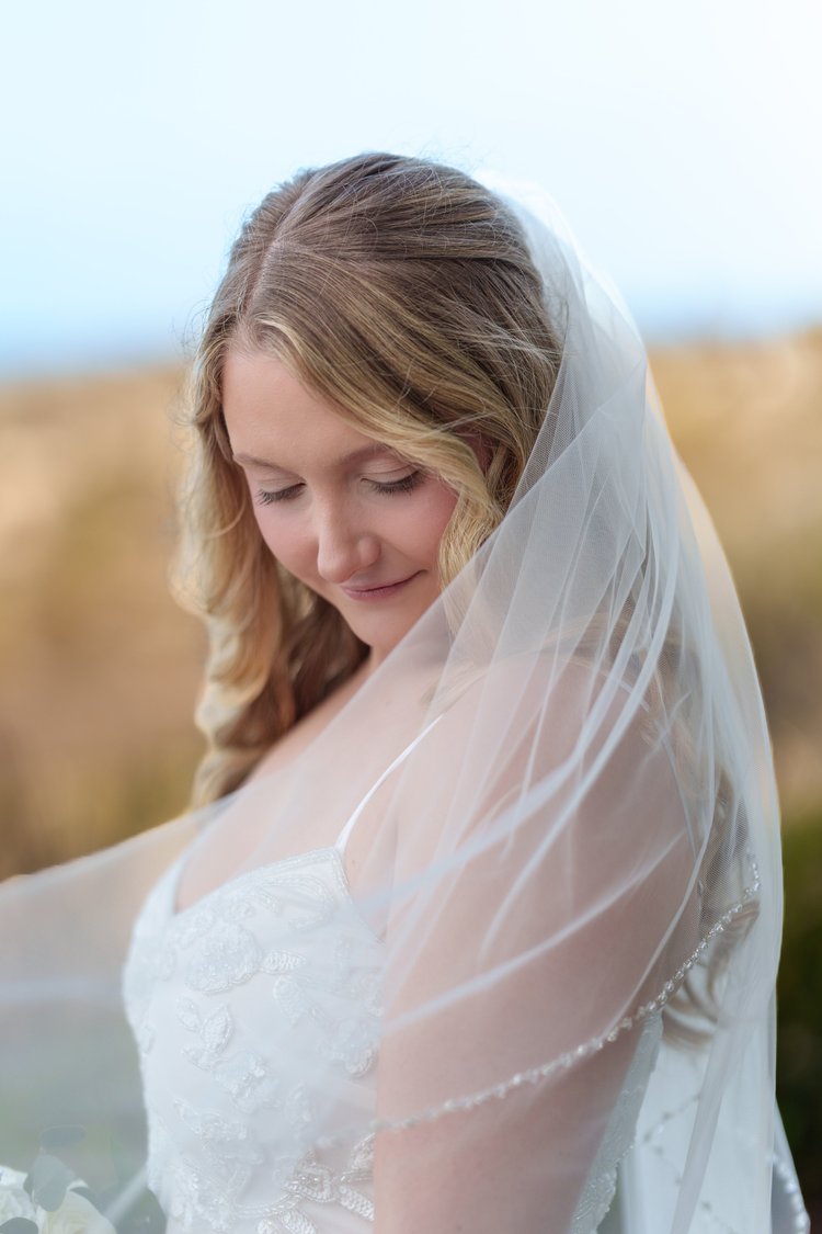 A portrait of a bride looking down and smiling, her veil draped over her shoulders in soft afternoon light.