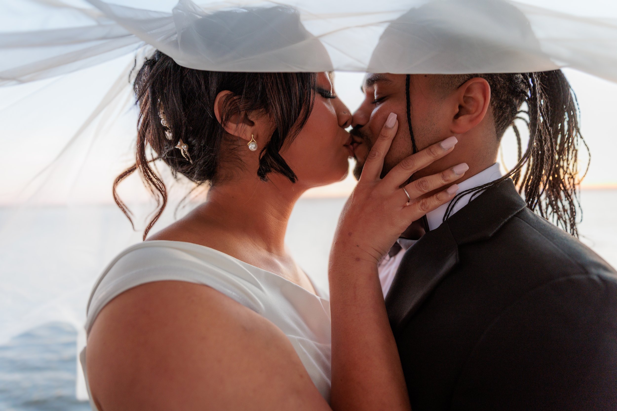 A couple kissing under a white umbrella by the water at sunset, dressed in wedding attire.