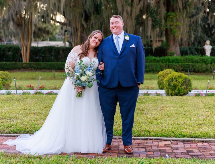 Bride and groom dressed in wedding attire, standing outdoors on a brick pathway with greenery and trees in the background. The bride wears a white gown with lace sleeves and holds a bouquet of white and blue flowers. The groom wears a blue suit with