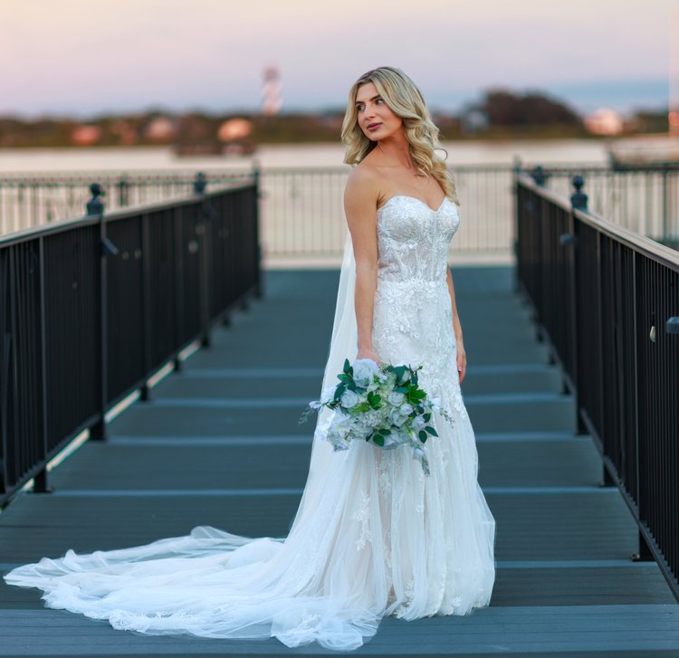 A bride in a white wedding gown holding a bouquet standing on a bridge at sunset
