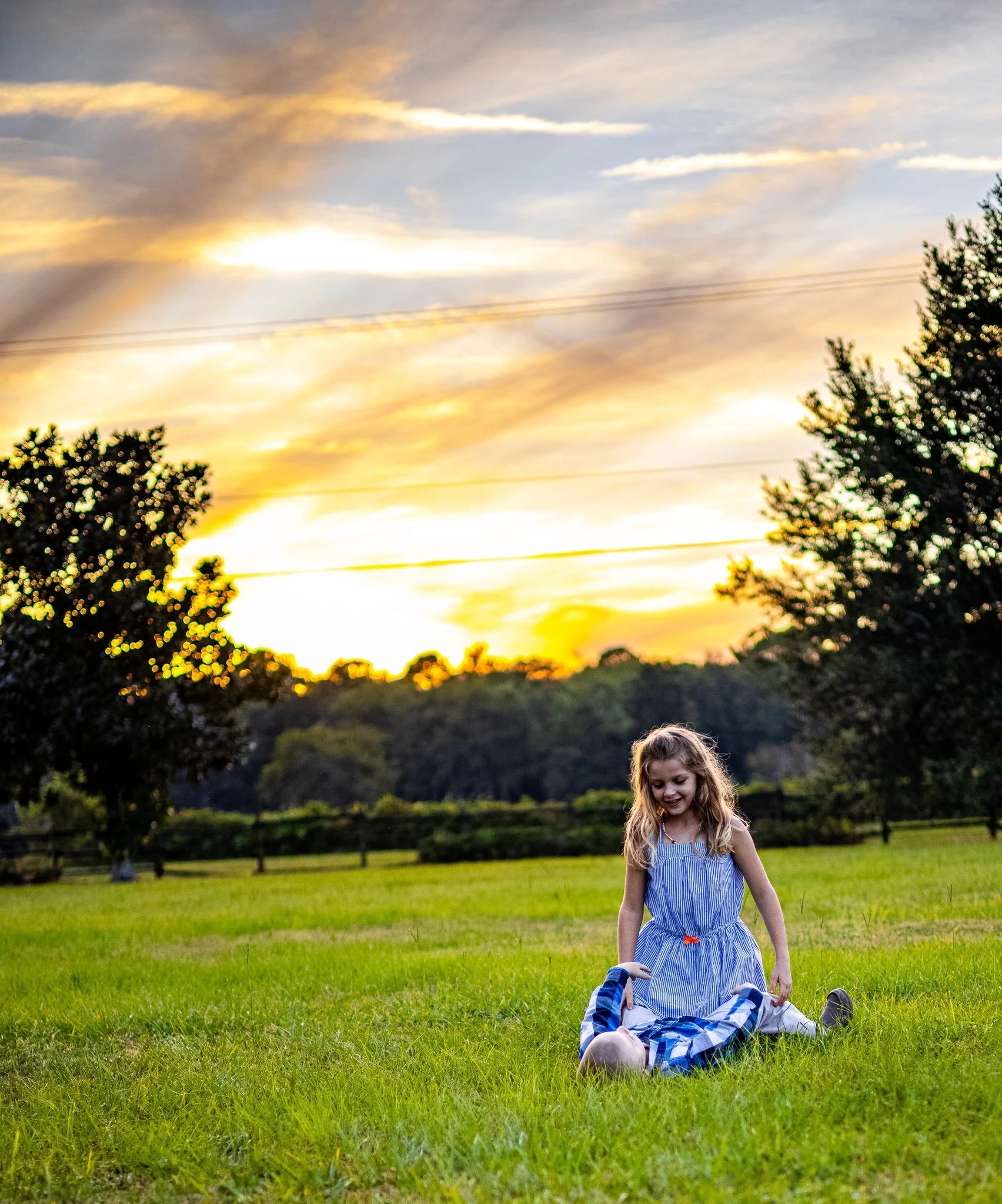 A young girl with long blonde hair in a blue dress playing on a grassy field at sunset.