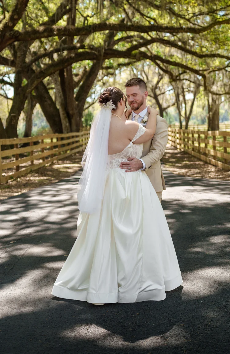 A bride and groom hold each other on a shaded path beneath an avenue of moss-draped oaks, her veil and full skirt catching the dappled light.