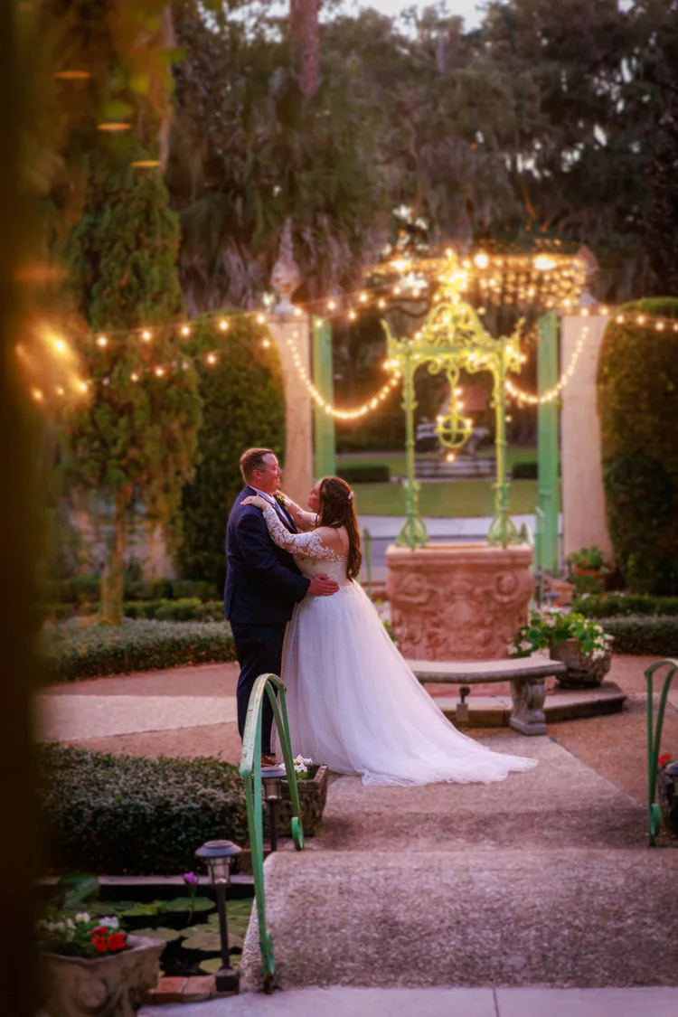 Bride and groom dancing outdoors at night under string lights, surrounded by trees and greenery, with a decorative water fountain in the background.