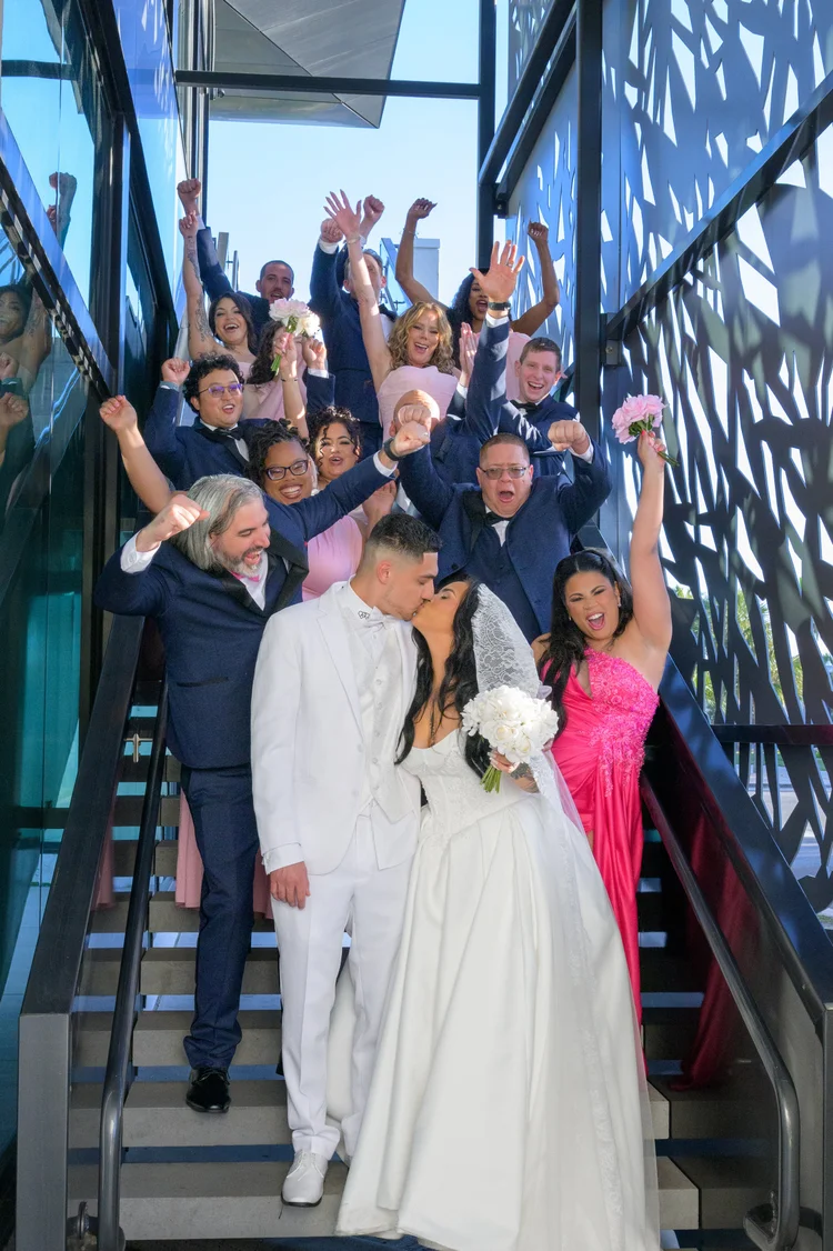 The wedding party cheers on a staircase behind the bride and groom, arms raised in celebration.