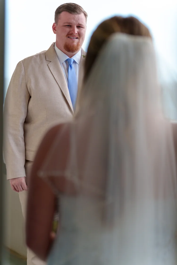A groom seeing his bride for the first time, smiling as she stands in the foreground with her veil down.