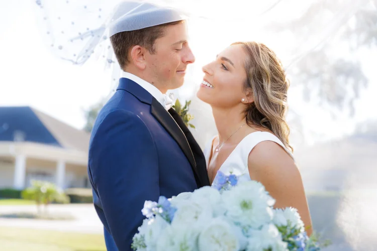 A bride and groom embrace outdoors on their wedding day, smiling at each other, with the bride holding a bouquet of white and light purple flowers.