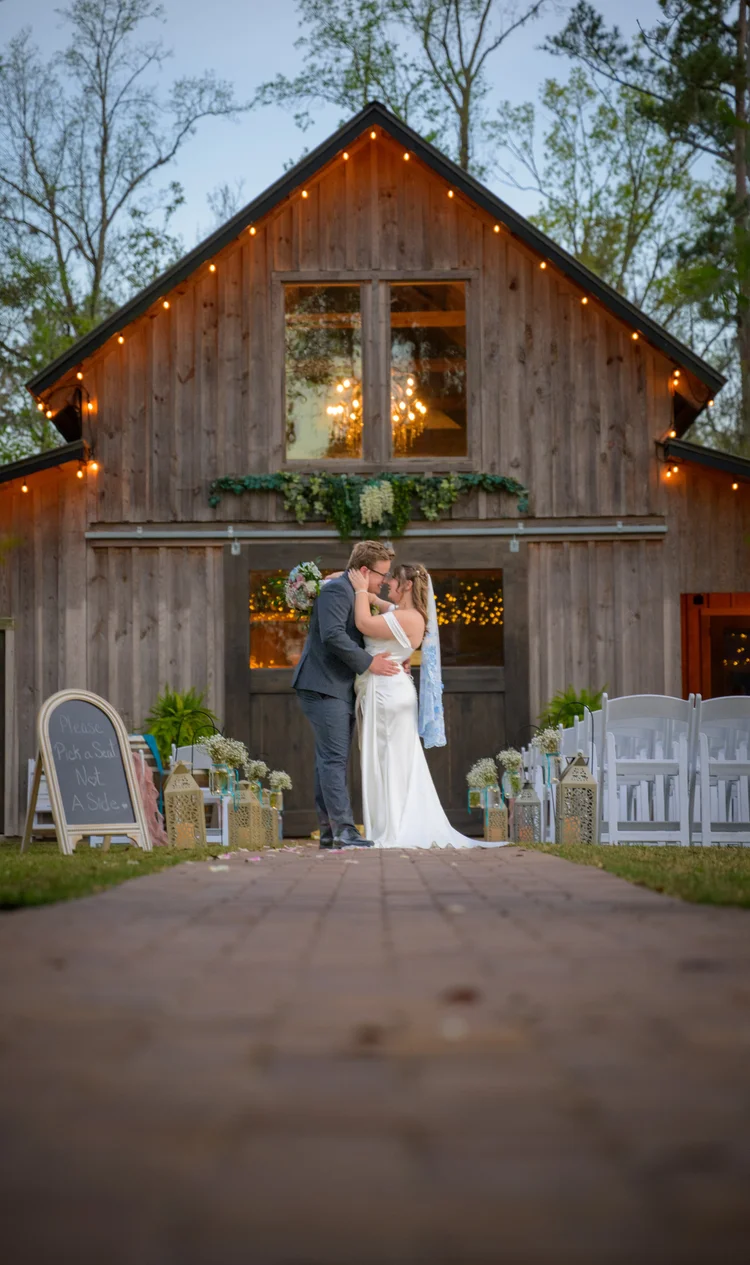 A bride and groom embrace in the open doorway of a warmly lit wooden barn at dusk, seen from the far end of the brick path.