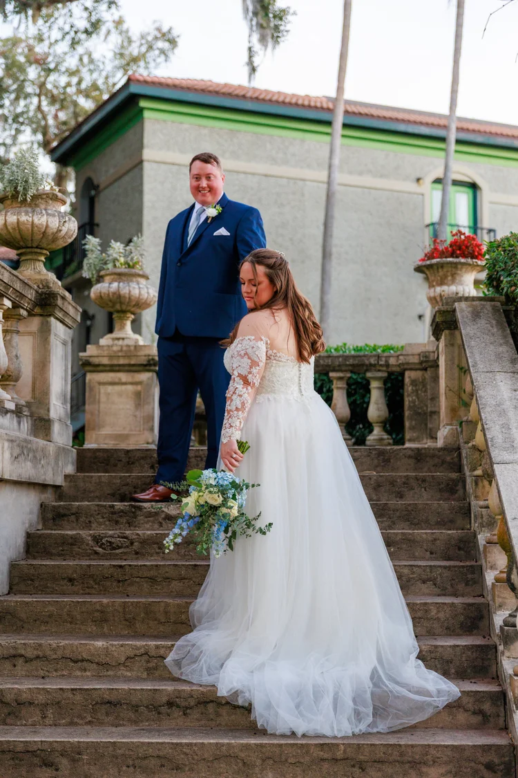 A bride in a white wedding gown holding a bouquet stands on stairs, looking down, while a groom in a blue suit stands above her, smiling and looking towards the camera.