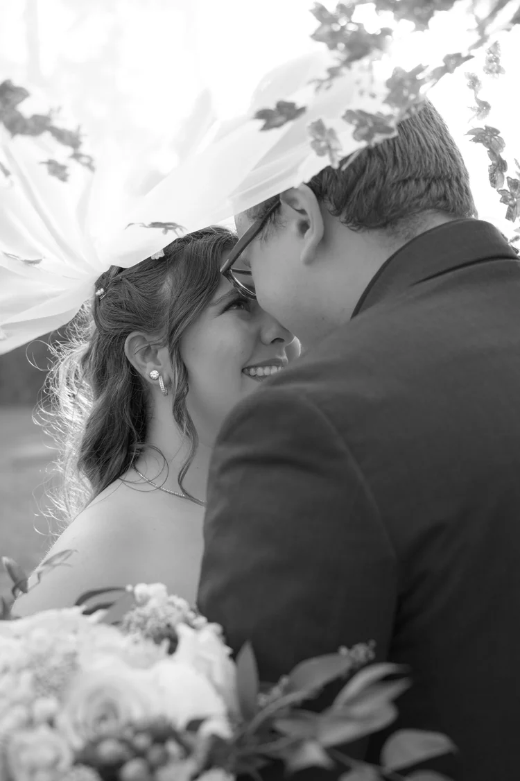 A black and white close-up of a bride and groom cheek to cheek beneath her veil, her bouquet just in frame.