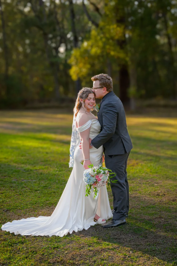A bride and groom stand close together on sunlit grass beneath tall trees, her gown trailing behind her.