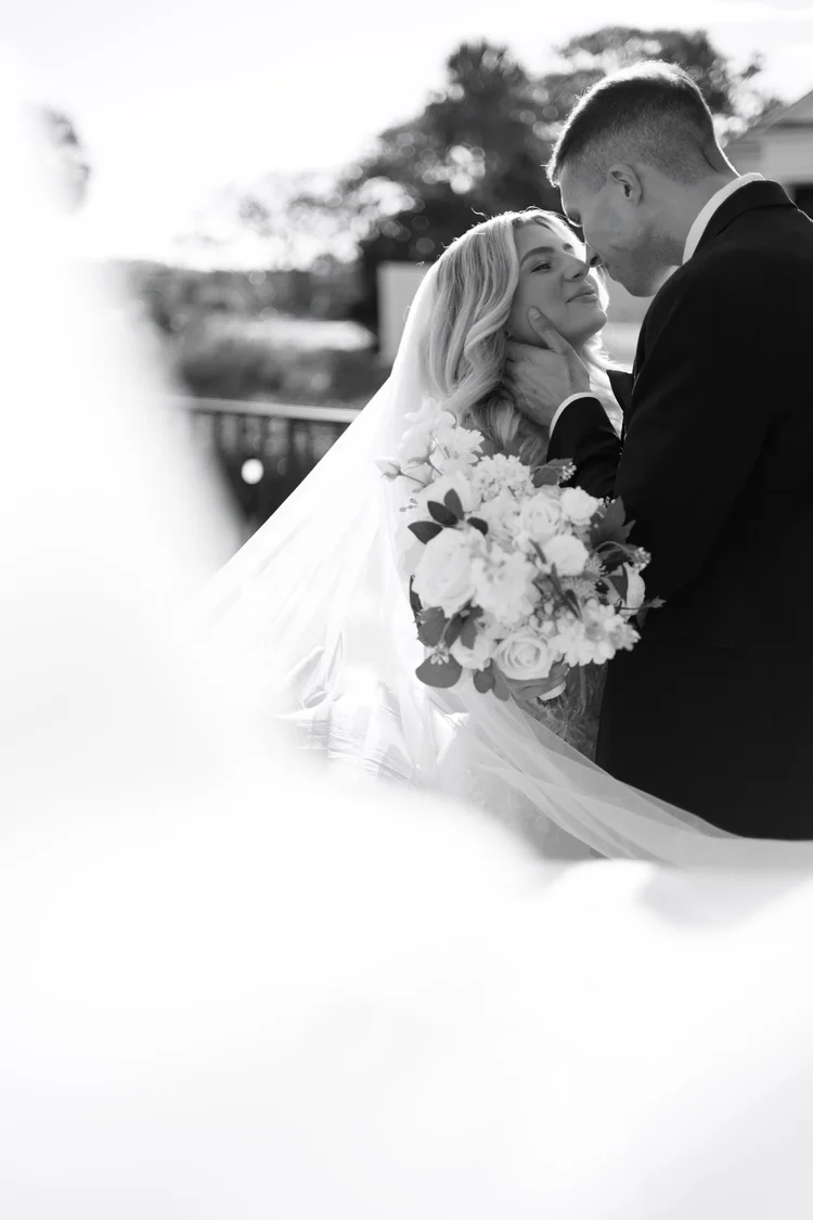 Black and white photo of a bride and groom sharing a romantic moment outside, with the groom holding the bride's face and the bride smiling, holding a bouquet of flowers.