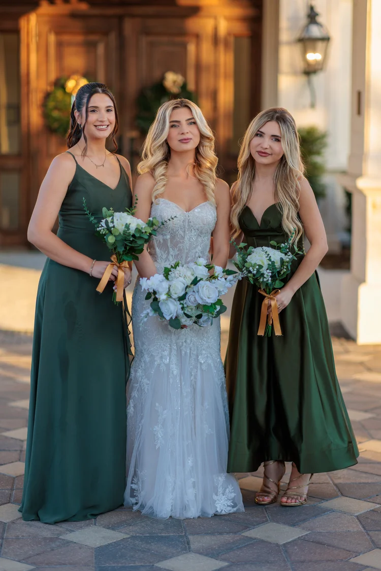 A bride in a fitted lace gown stands between two bridesmaids in deep green dresses, all three holding white and greenery bouquets in front of tall wooden doors.
