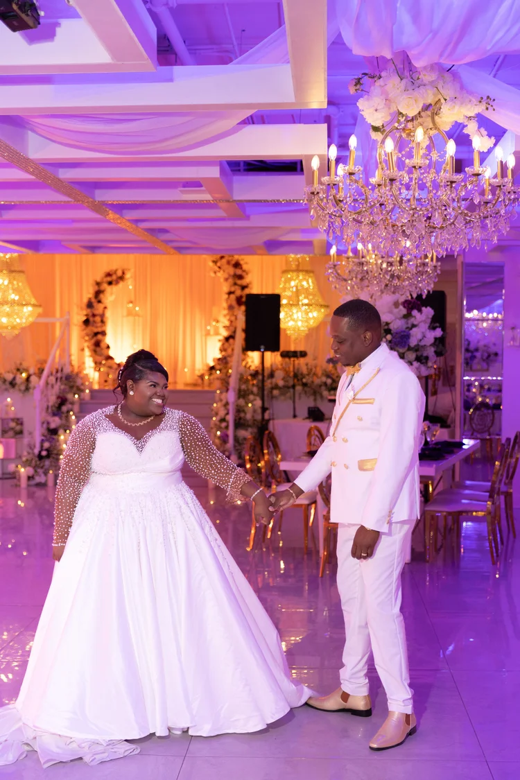 A bride and groom face each other on the dance floor of a reception room lit in warm amber and violet, chandeliers overhead and a floral arch behind them.
