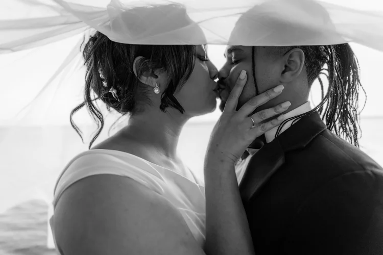 A black and white photo of a couple kissing, with the woman touching the man's face, under a large umbrella at the beach.