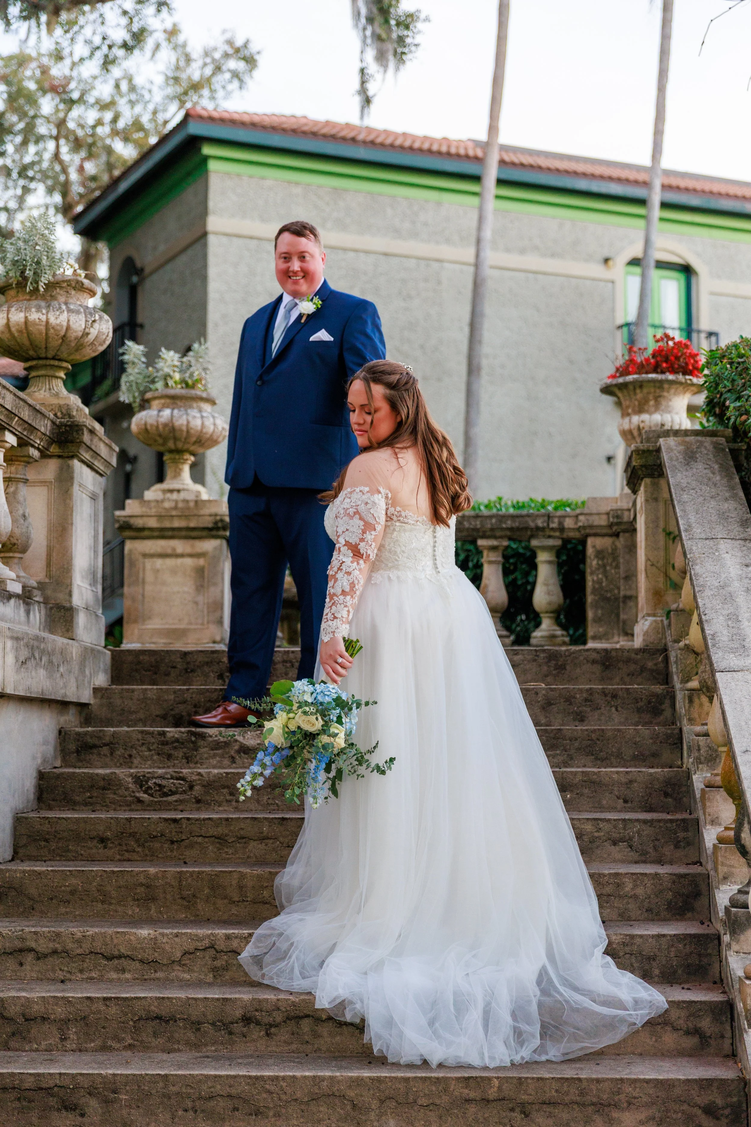 A bride in a white wedding gown holding a bouquet stands on stairs, looking down, while a groom in a blue suit stands above her, smiling and looking towards the camera.
