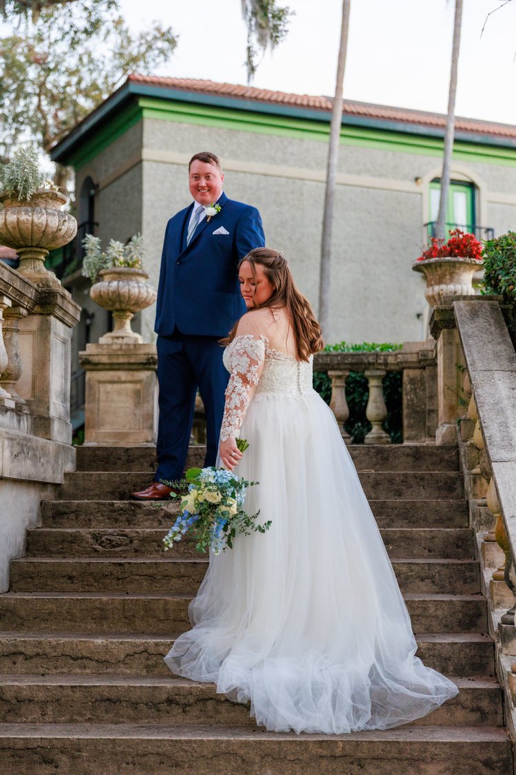 A bride in a white wedding gown holding a bouquet stands on stairs, looking down, while a groom in a blue suit stands above her, smiling and looking towards the camera.