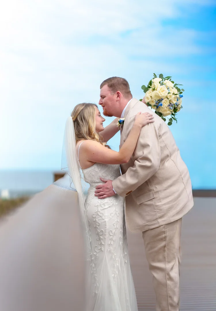 A bride and groom hold each other on an oceanfront deck under a bright blue sky, her veil lifting in the wind and a yellow and white bouquet in his hand.