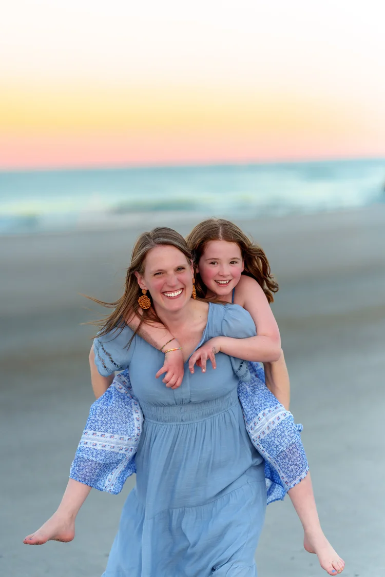 A mother in a blue dress holds her laughing daughter on the beach at sunset.