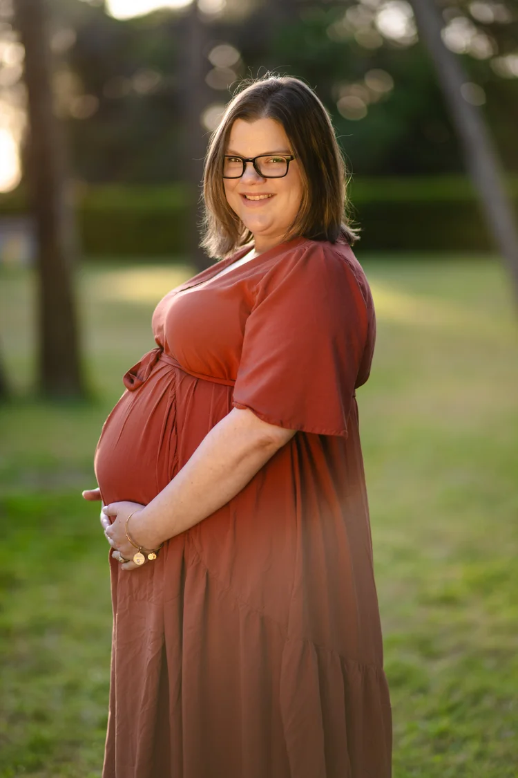 An expectant mother in a rust-coloured dress rests her hands on her belly in a park at golden hour.