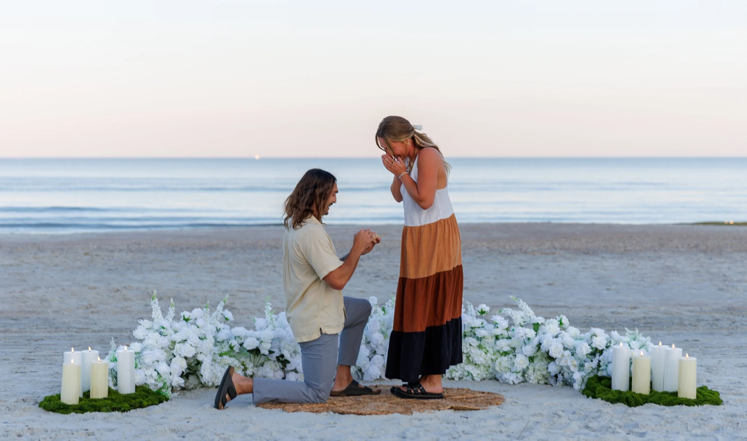 A man proposes to a woman on a beach during sunset with flowers and candles around them.