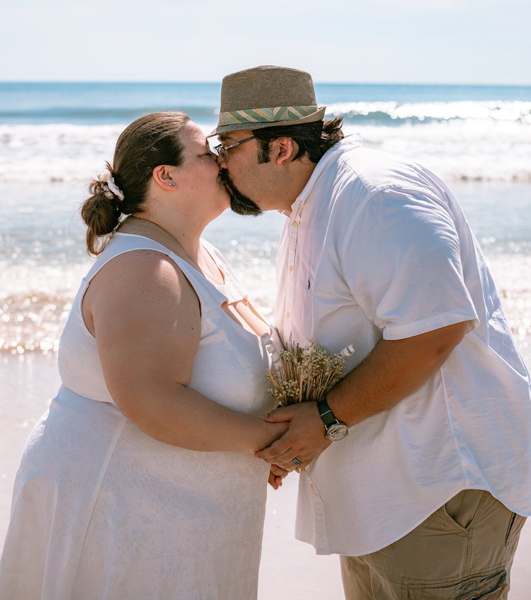 A couple kiss on the beach in white wedding attire, the surf breaking behind them and a small bouquet held at her wrist.