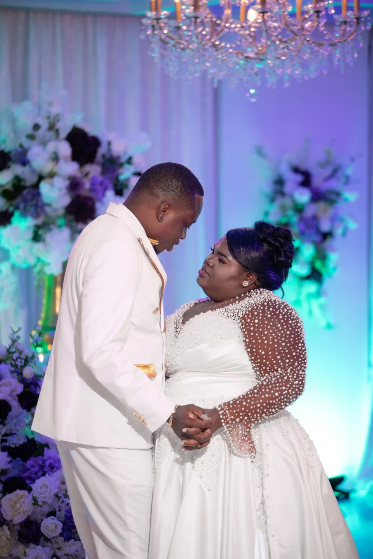 A newlywed couple in matching white attire hold each other close during their first dance, framed by a floral backdrop and a crystal chandelier.
