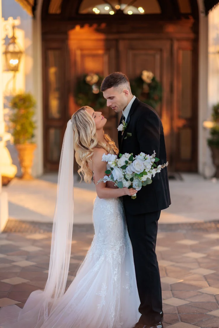 A bride and groom lean into each other on a paved courtyard at golden hour, her veil trailing behind her and a white bouquet held between them.