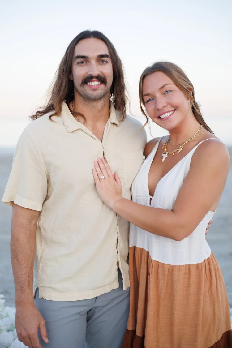 An engaged couple stand close together on the beach at sunset, the woman resting a hand on his chest.