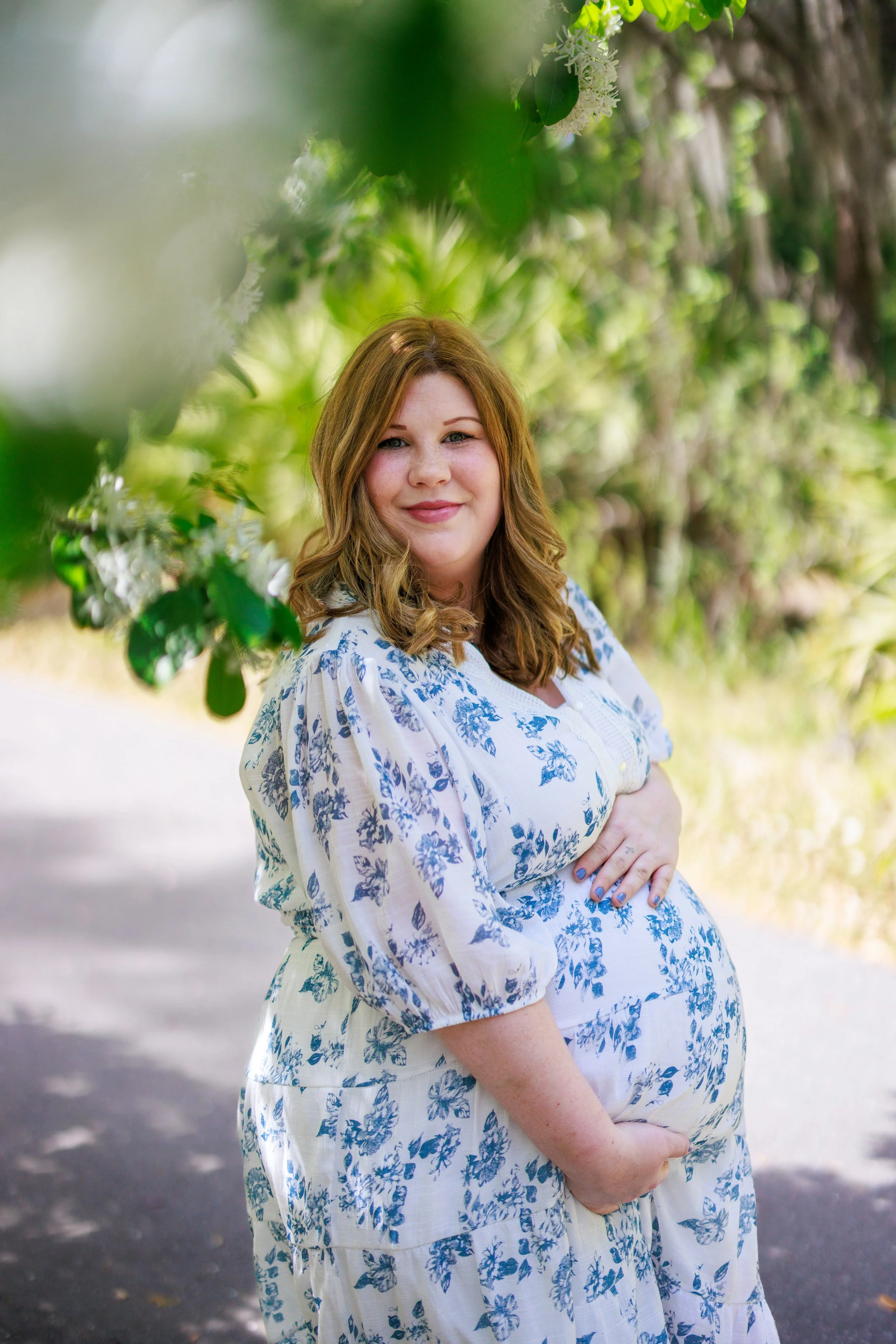 A pregnant woman with wavy, shoulder-length hair wearing a white dress with blue floral patterns, smiling outdoors among greenery.
