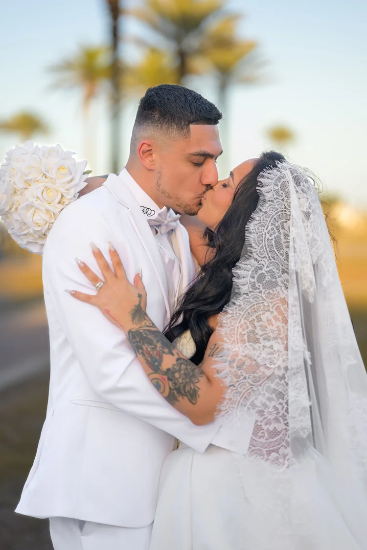 A bride and groom hold each other and kiss at sunset, palm trees blurred in the background and her veil lifted around them.