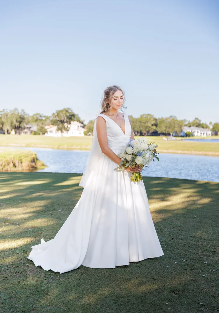 A bride in a white wedding dress holding a bouquet of white and blue flowers on a grassy area near a lake with houses and trees in the background, under a clear blue sky.
