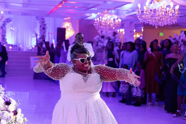 A bride throws her arms wide in celebration on the dance floor, guests gathered and cheering behind her under purple reception lighting.