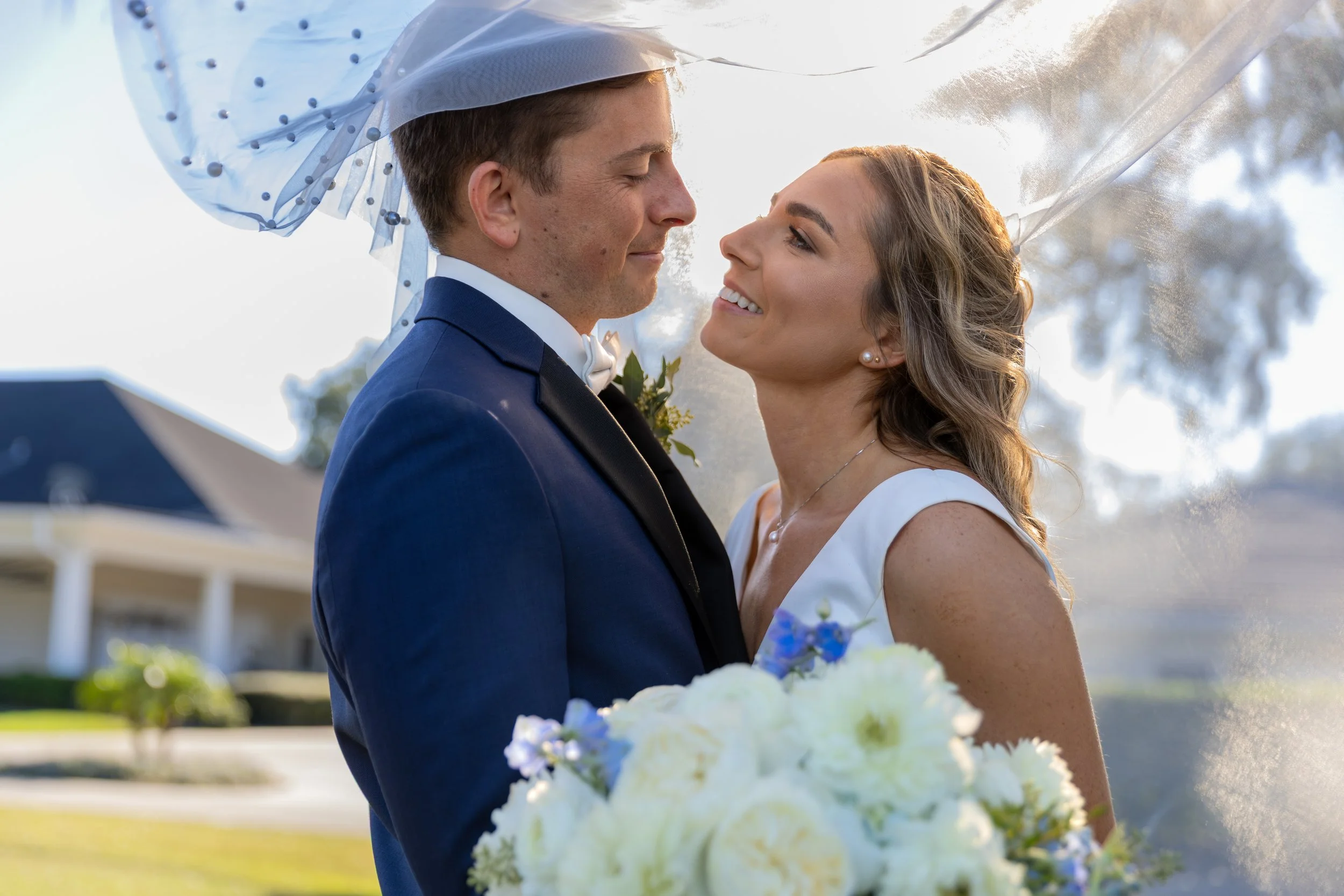 A bride and groom facing each other outdoors, with trees in the background. The groom is wearing a navy suit with a white shirt and bow tie, while the bride is wearing a white dress and holding a bouquet of white and purple flowers. They are smiling 