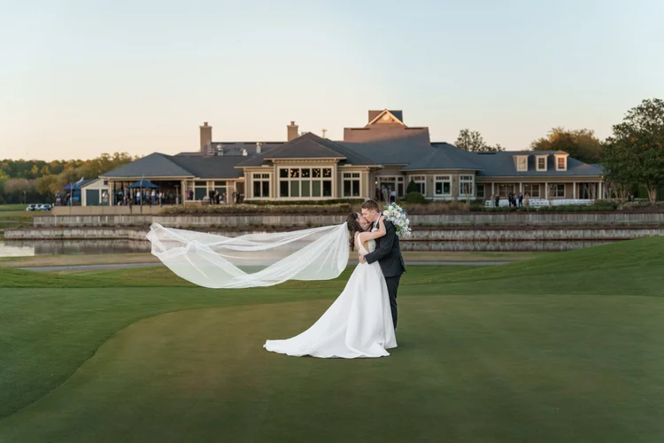 A bride and groom embrace on a manicured lawn at sunset with a clubhouse and water behind them, her veil stretched out across the grass.