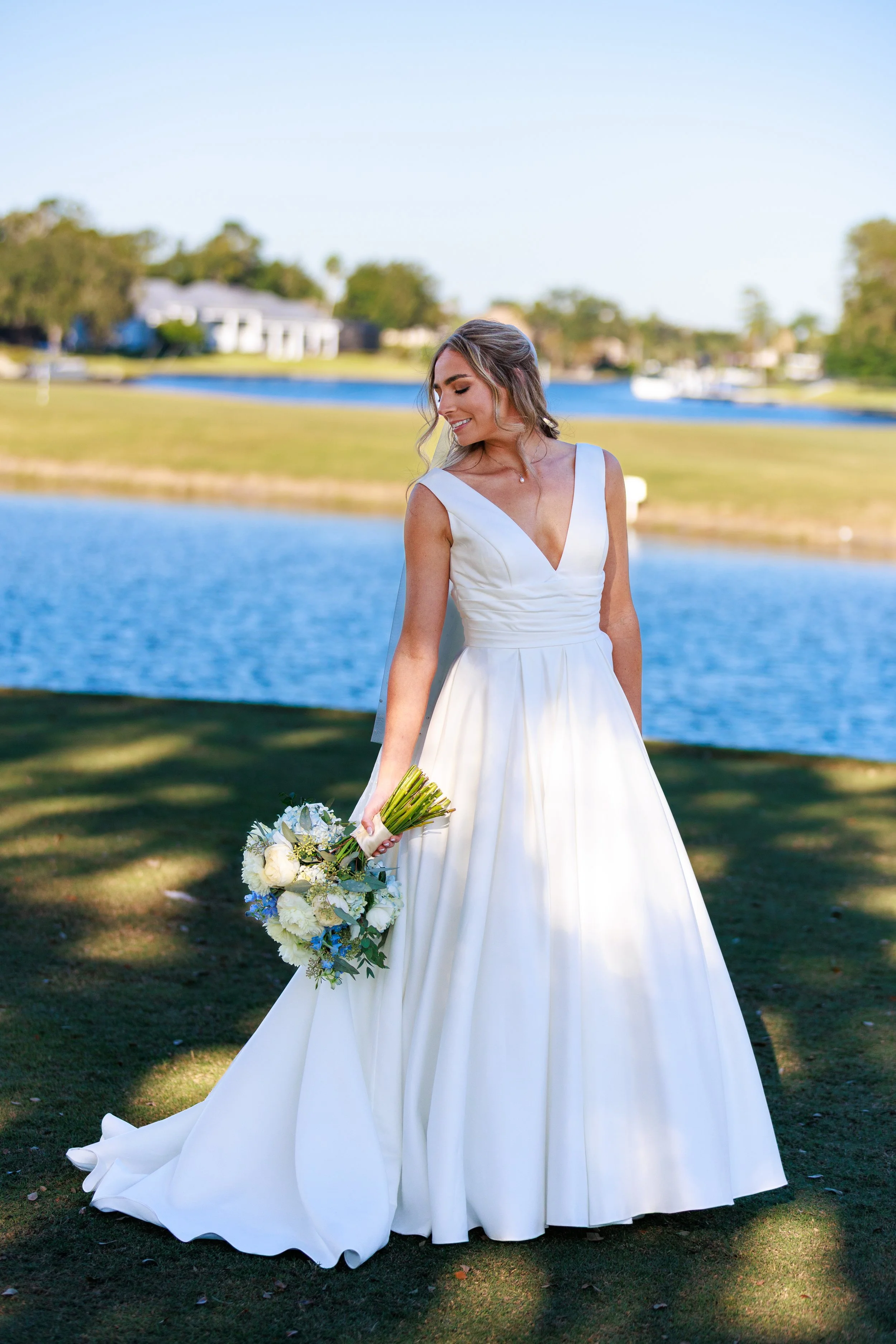 A smiling woman in a white wedding dress holding a bouquet outdoors near a body of water.