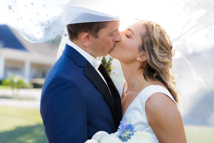 A bride and groom sharing a kiss outdoors on their wedding day, with the bride holding a bouquet of flowers.