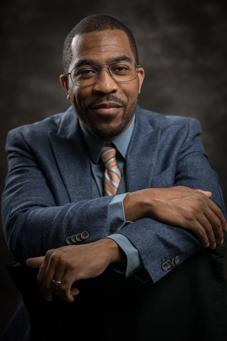A studio headshot of a man in a grey suit and patterned tie, arms folded, against a dark backdrop.